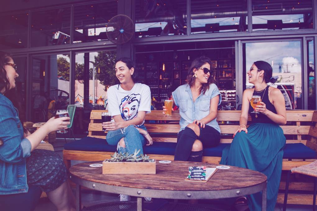 Four women sitting on bench in storefront while drinking alcoholic beverages.