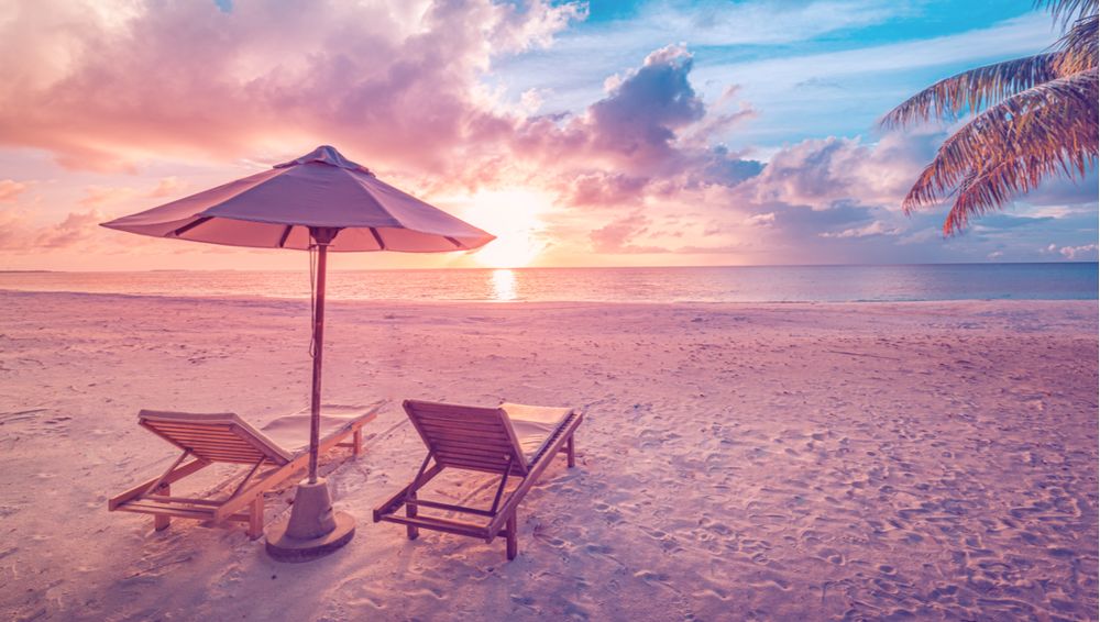 Tropical sunset scenery with two loungers on beach and umbrella under palm tree.