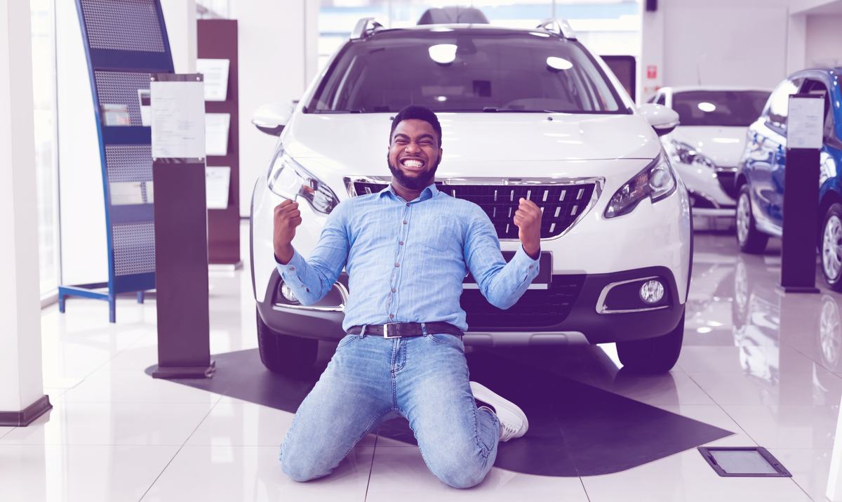 Excited Black Guy Shaking Fists Standing On Knees Near Auto In Dealership Store.