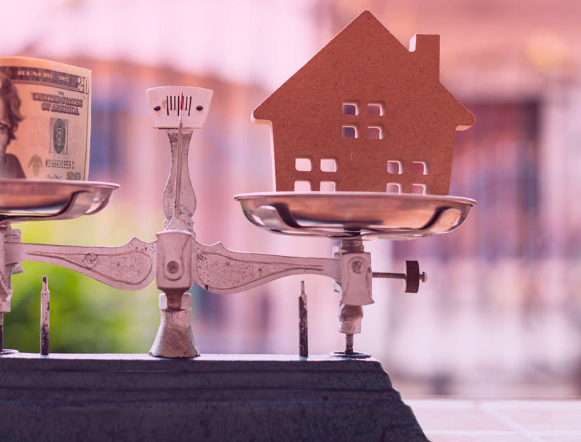 How-Much-Should-You-Have-In-the-Bank-To-Buy-A-Home House model and money on weight scale with out of focus home.