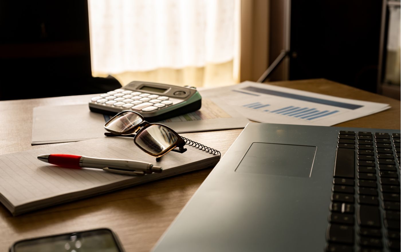 Work table at home with pen, papers, bills, charts, glasses, computer and calculator.