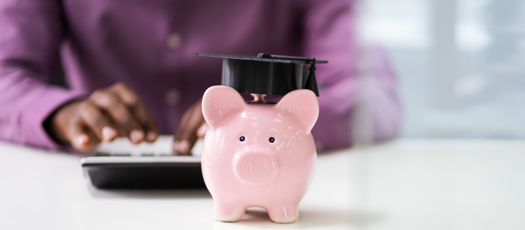 Businessperson's hand using calculator beside piggybank with graduation hat.